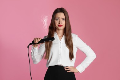Image of Upset young woman with flattening iron on light pink background. Hair damage