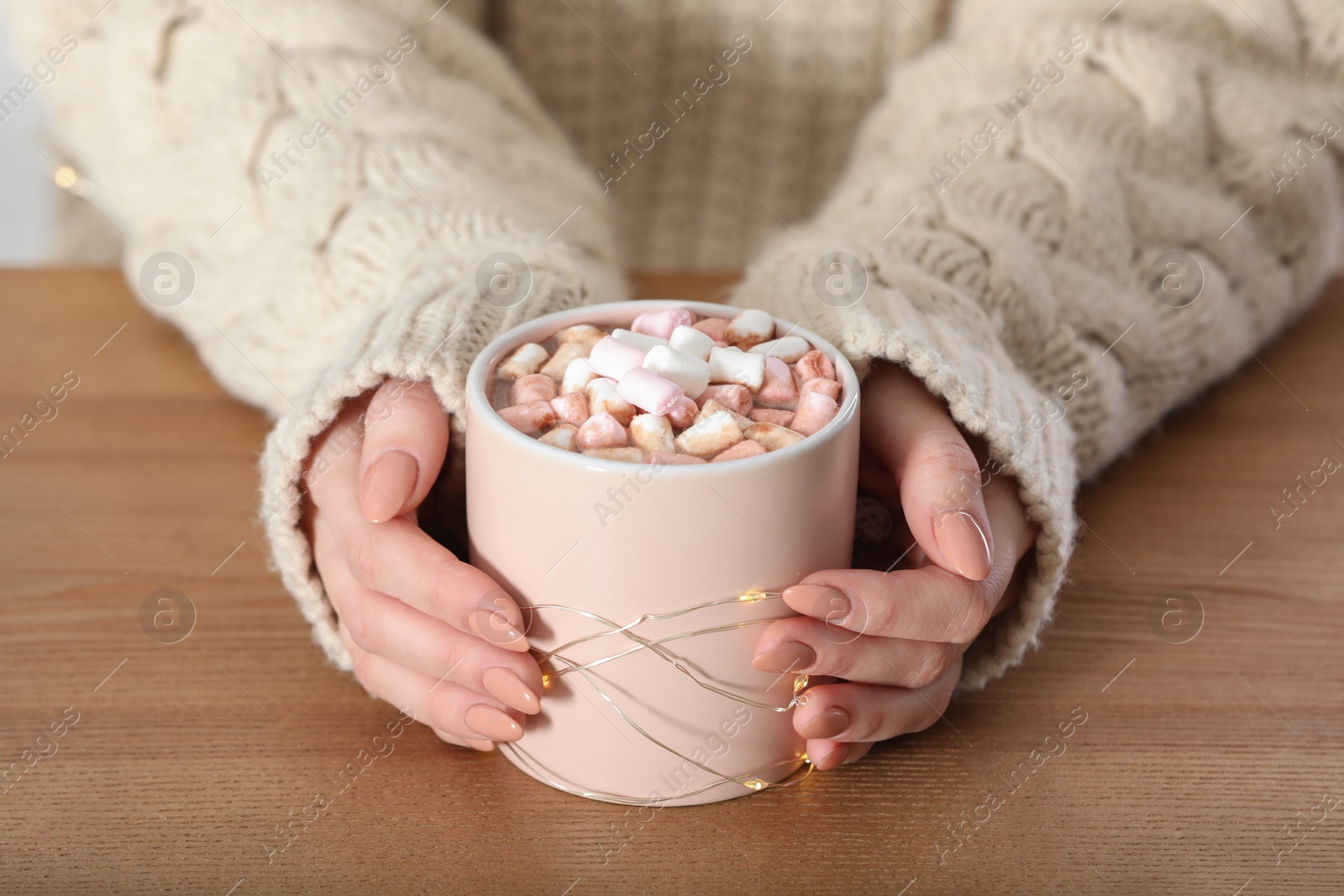 Photo of Woman in sweater holding cup of aromatic cacao with marshmallows on wooden table, closeup