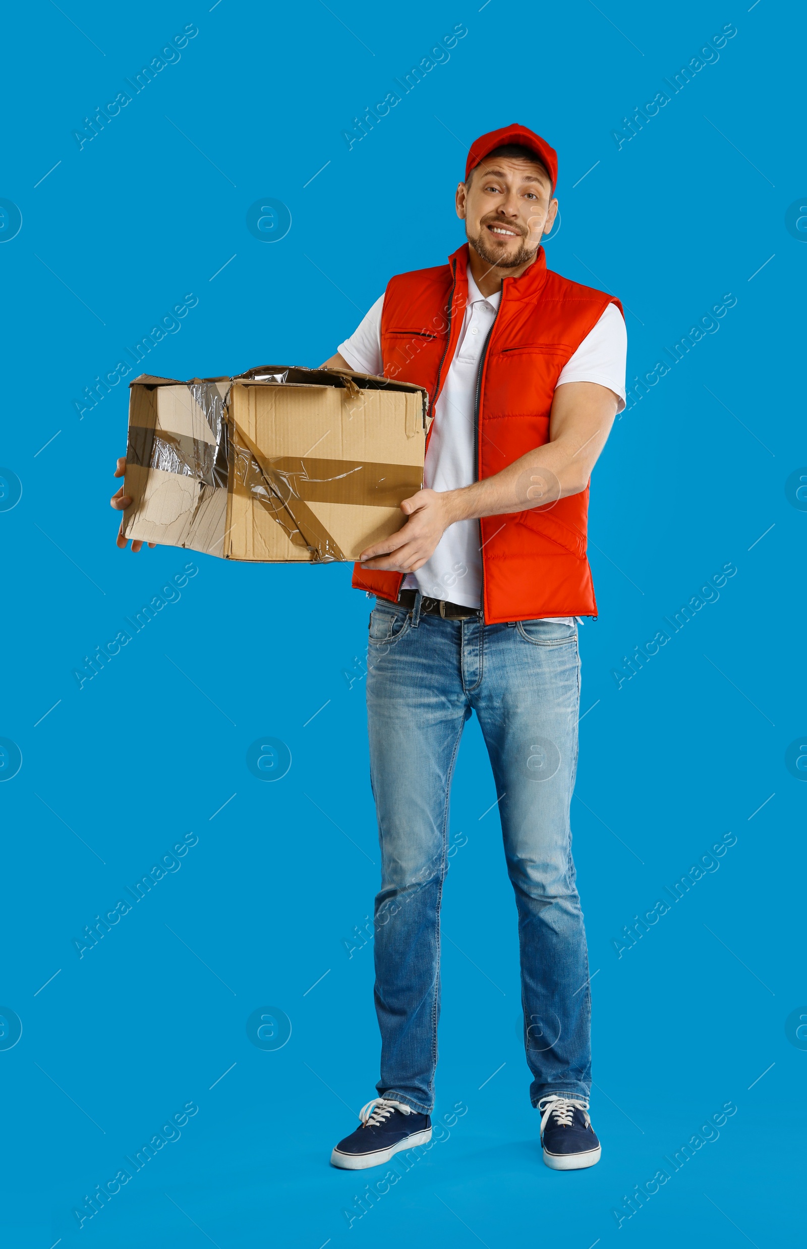 Photo of Emotional courier with damaged cardboard box on blue background. Poor quality delivery service