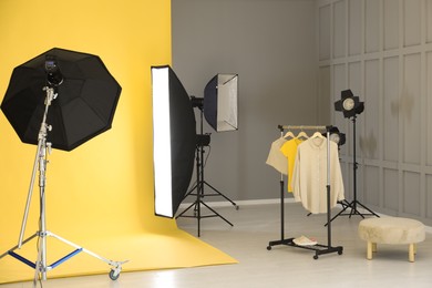 Photo of Yellow photo background, rack with clothes and professional lighting equipment in modern studio