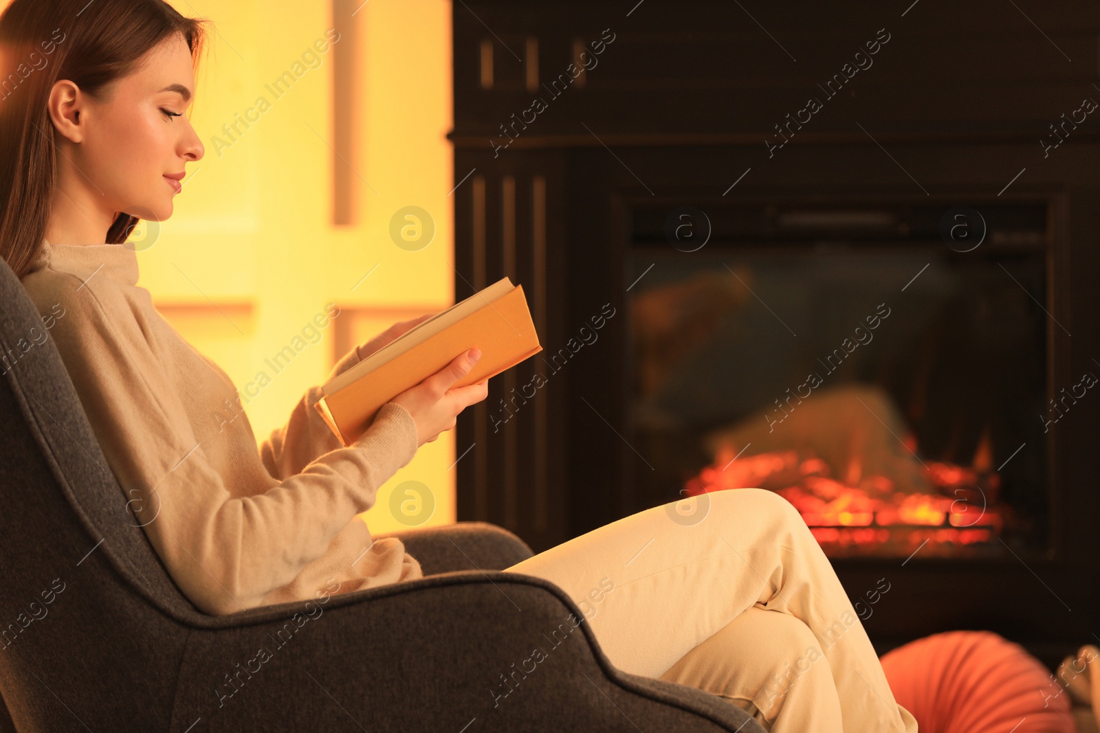 Photo of Young woman reading book near fireplace indoors. Cozy atmosphere