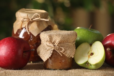 Photo of Delicious apple jam and fresh fruits on burlap against blurred background, closeup