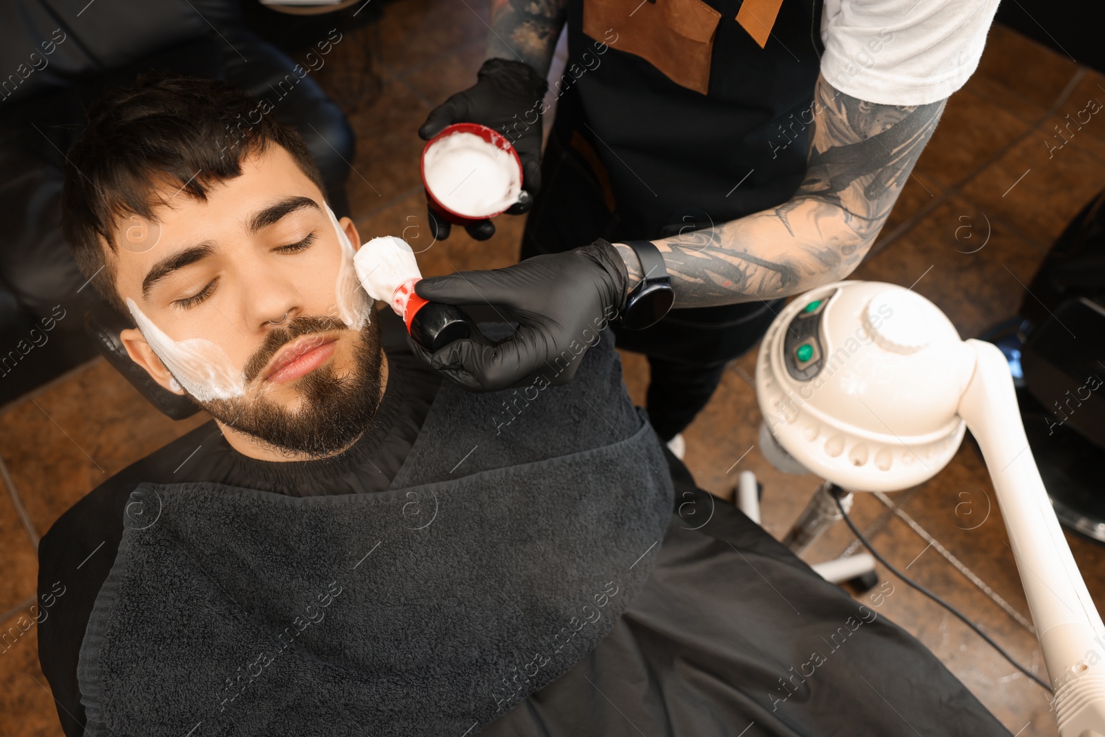 Photo of Professional hairdresser applying shaving foam onto client's beard in barbershop, closeup