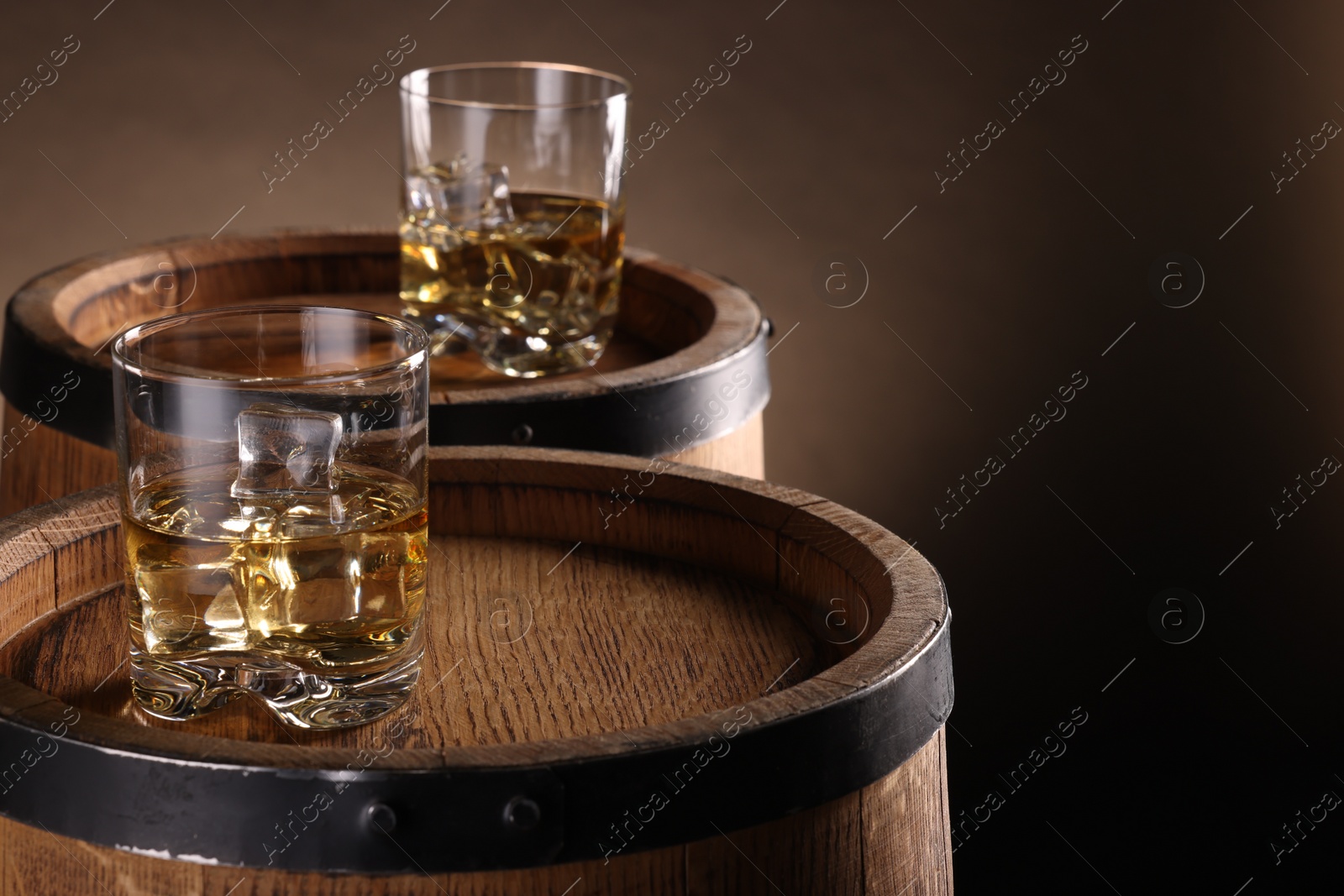 Photo of Whiskey with ice cubes in glasses on wooden barrels against brown background, closeup. Space for text