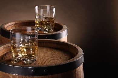 Photo of Whiskey with ice cubes in glasses on wooden barrels against brown background, closeup. Space for text