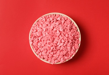 Photo of Bright heart shaped sprinkles in white bowl on red background, top view