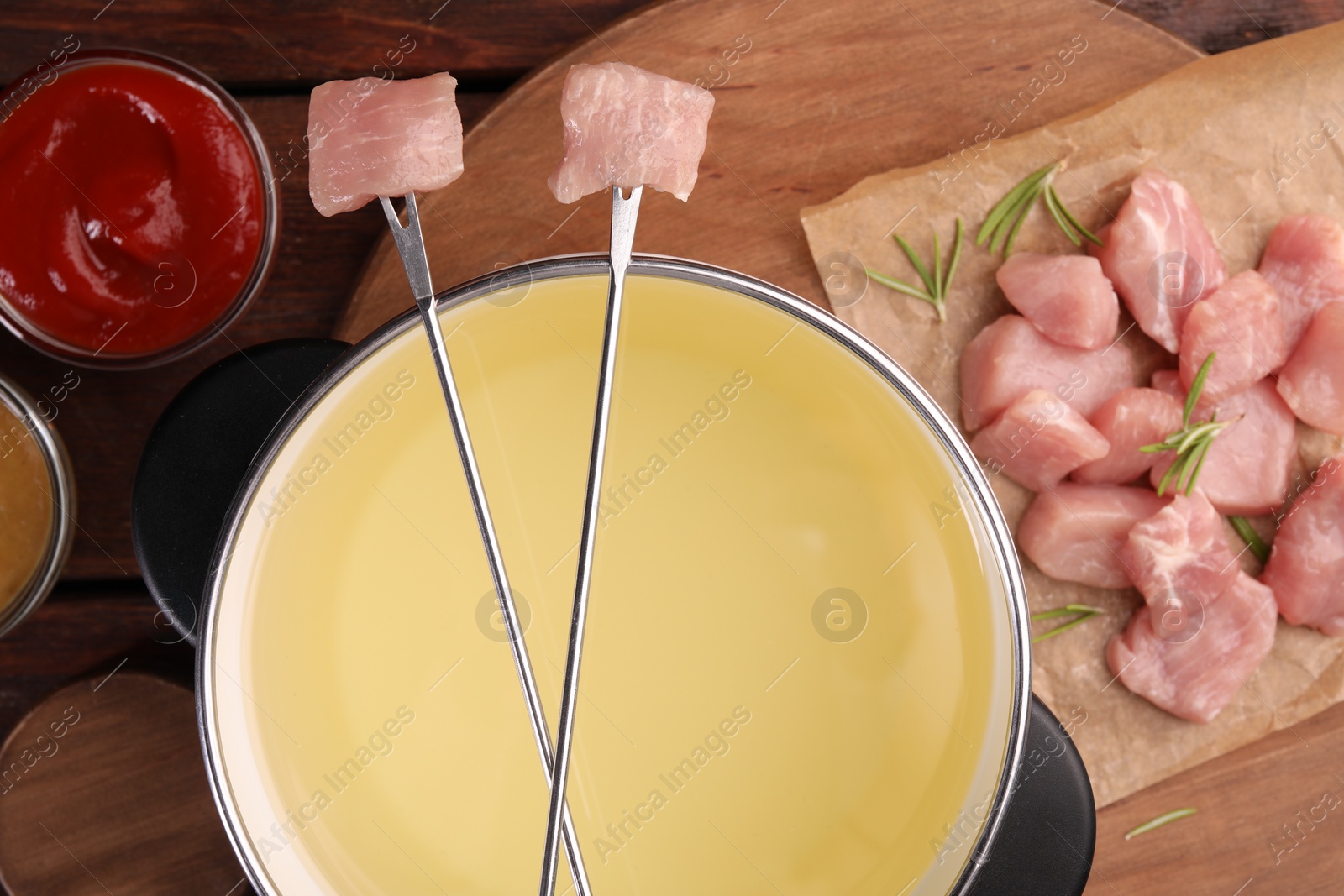 Photo of Fondue pot, forks with pieces of raw meat and sauces on wooden table, flat lay