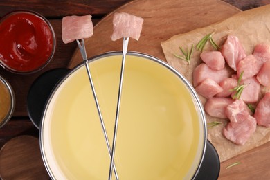 Fondue pot, forks with pieces of raw meat and sauces on wooden table, flat lay