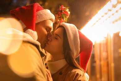 Photo of Happy couple in Santa hats kissing under mistletoe bunch outdoors, bokeh effect