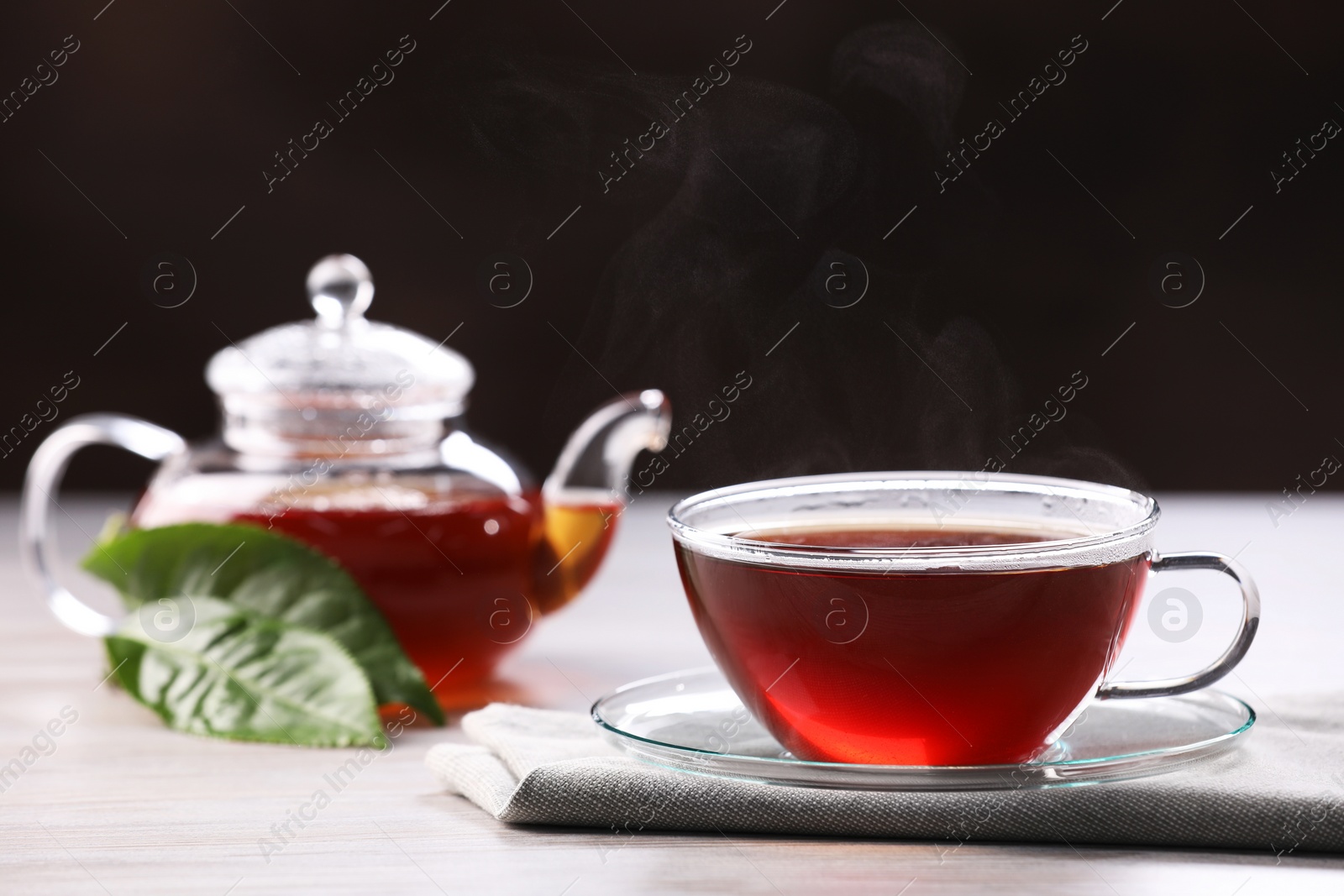 Photo of Aromatic hot tea in glass cup, teapot and leaves on white wooden table against black background. Space for text