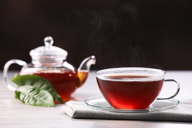 Photo of Aromatic hot tea in glass cup, teapot and leaves on white wooden table against black background. Space for text