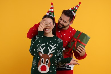 Couple in Christmas sweaters. Young man surprising his woman with gift on orange background