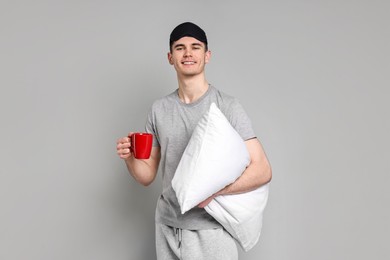 Man in pyjama holding pillow and cup of drink on grey background