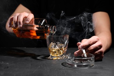 Photo of Alcohol addiction. Man with smoldering cigarette pouring whiskey into glass at dark textured table, closeup