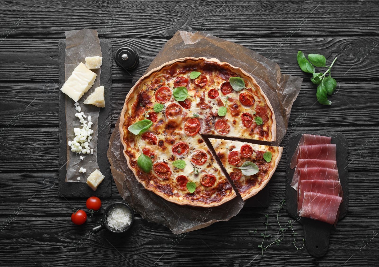 Photo of Cut delicious homemade quiche with prosciutto and ingredients on black wooden table, flat lay