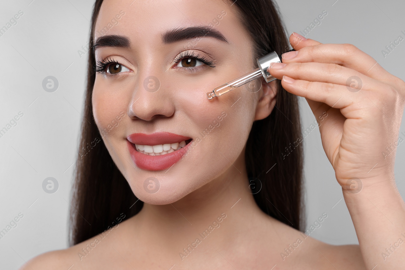 Photo of Happy young woman applying essential oil onto face on light grey background, closeup