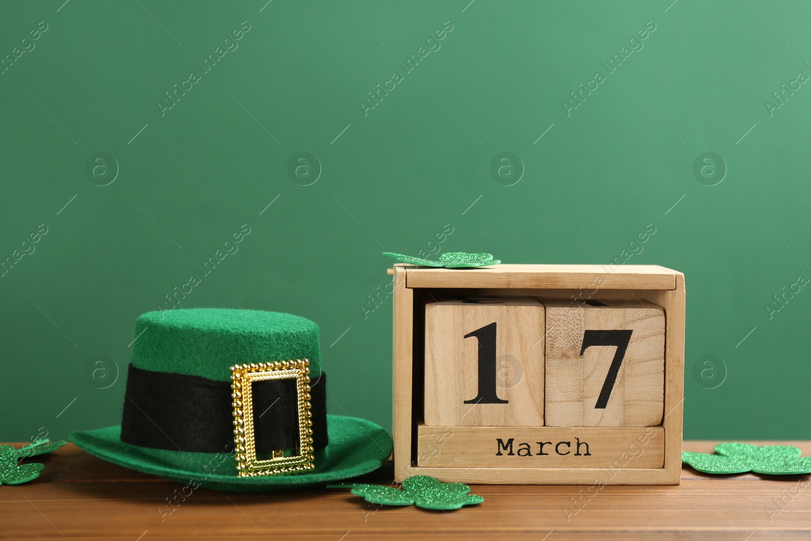 Photo of Leprechaun hat, clover leaves and block calendar on wooden table against green background, space for text. St Patrick's Day celebration