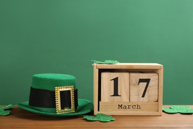 Leprechaun hat, clover leaves and block calendar on wooden table against green background, space for text. St Patrick's Day celebration