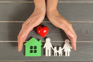 Woman holding hands near figures of house, red heart and family on grey wooden background, top view