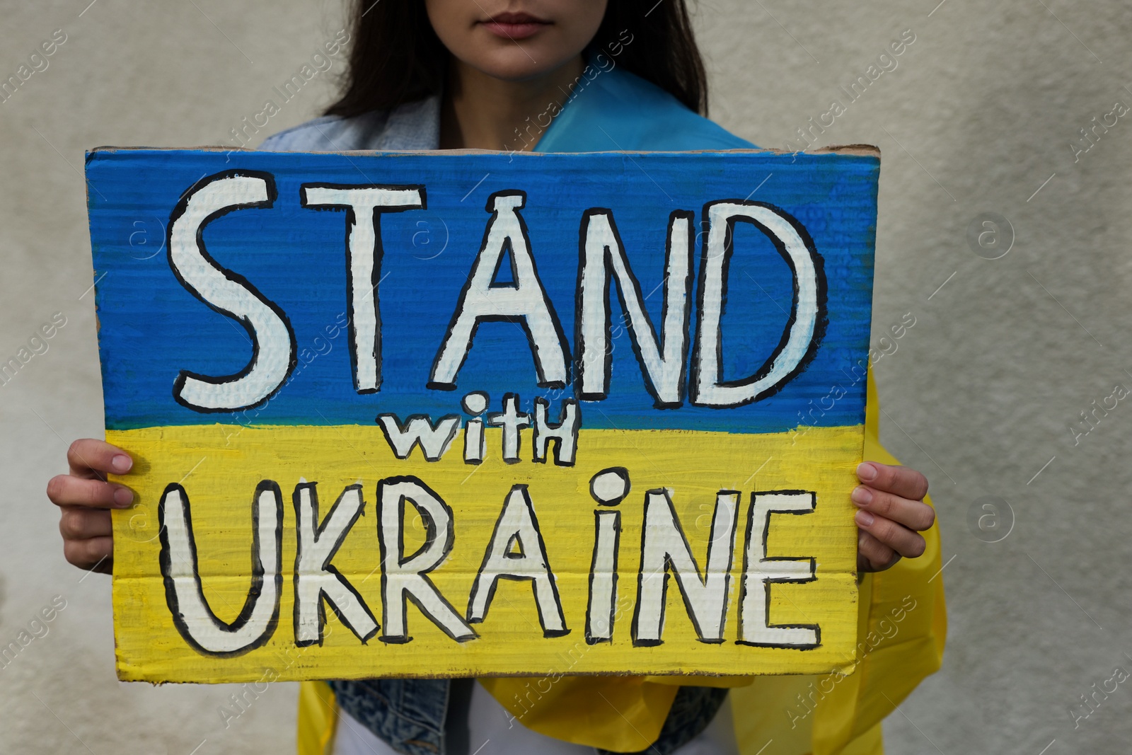 Photo of Young woman holding poster in colors of national flag and words Stand with Ukraine near light wall, closeup