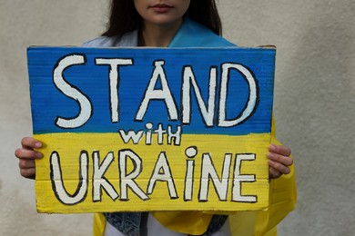 Young woman holding poster in colors of national flag and words Stand with Ukraine near light wall, closeup