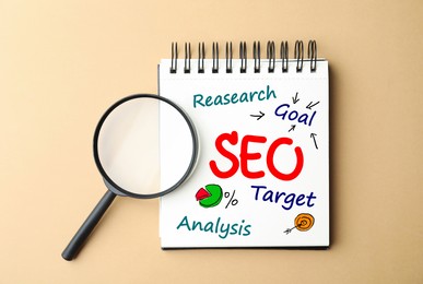 Search engine optimization. Notebook with abbreviation SEO and magnifying glass on beige background, top view