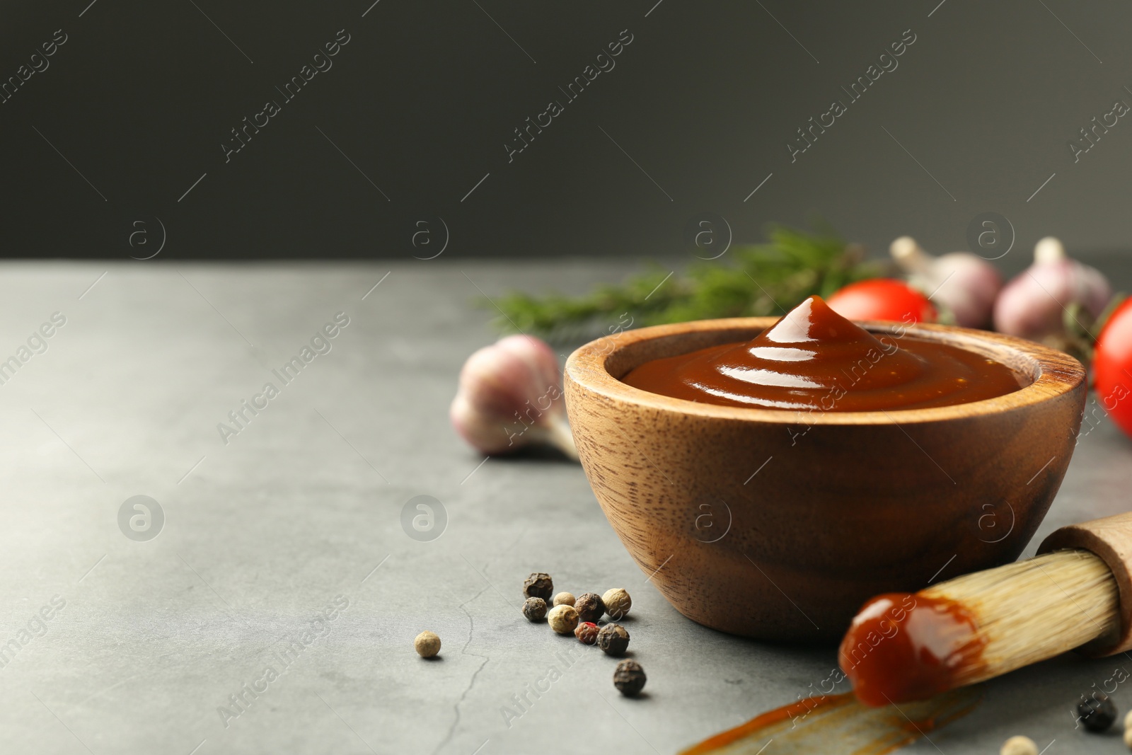 Photo of Tasty barbeque sauce in bowl, brush and peppercorns on grey textured table, closeup. Space for text