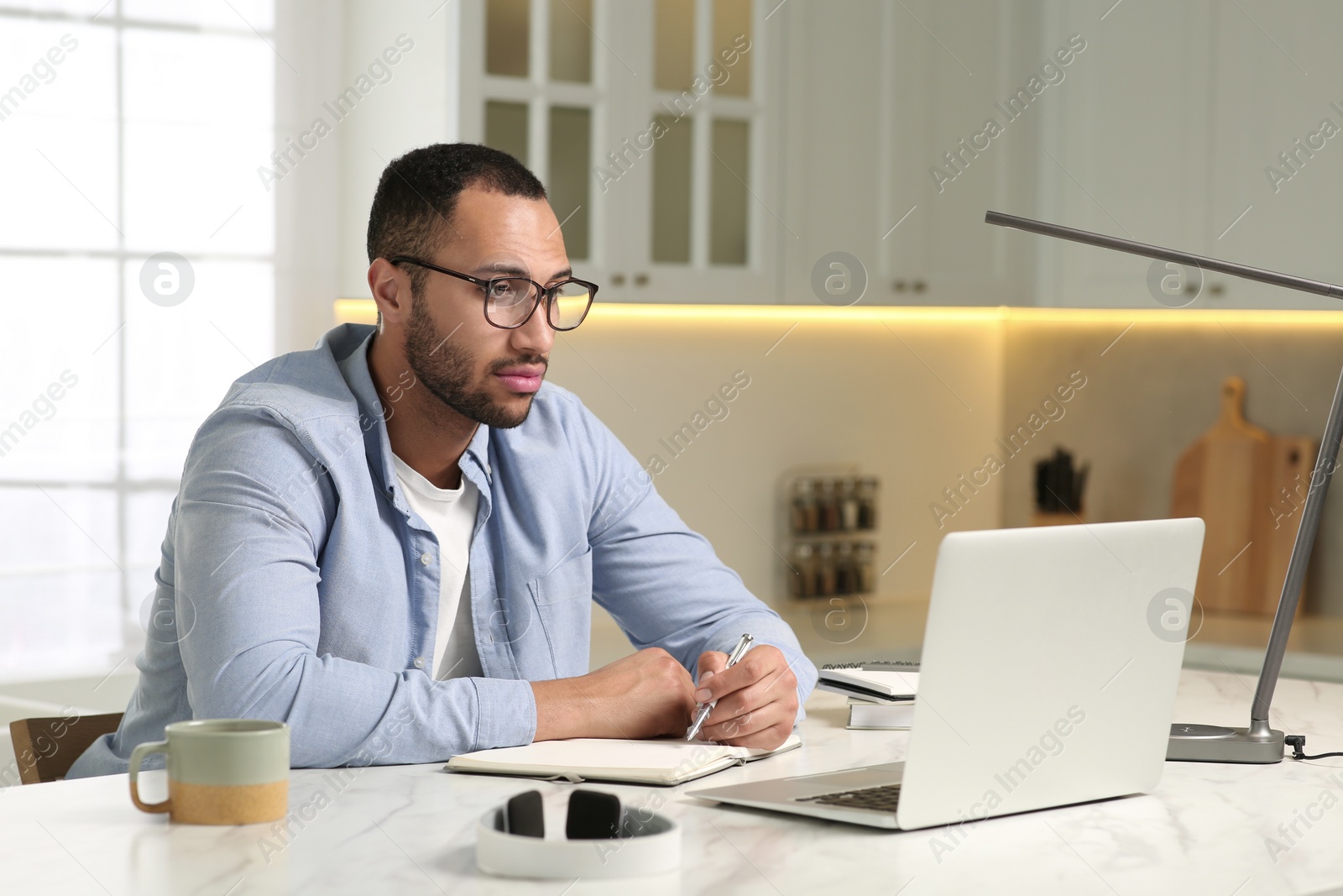 Photo of Young man with laptop writing in notebook at desk in kitchen. Home office