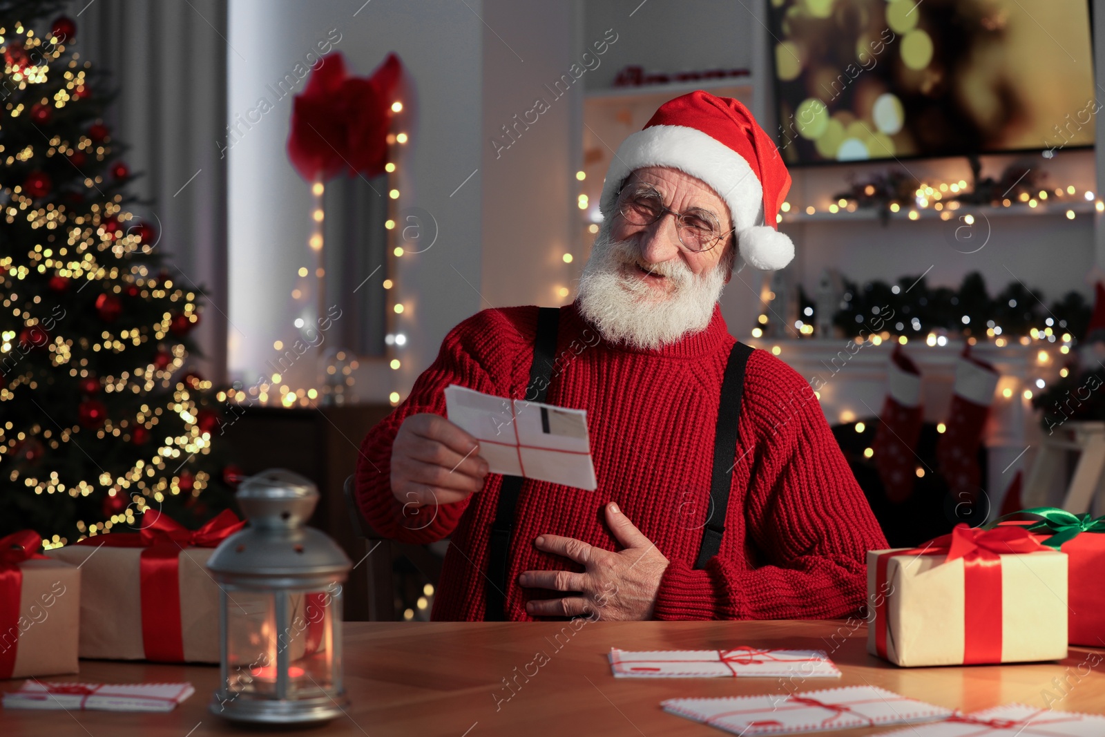 Photo of Santa Claus reading letter and laughing at his workplace in room decorated for Christmas