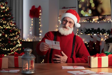 Santa Claus reading letter and laughing at his workplace in room decorated for Christmas