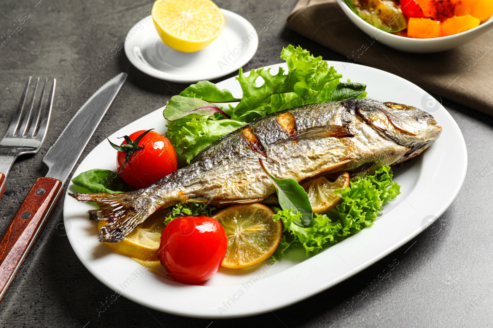 Photo of Delicious roasted fish with lemon and vegetables on dark grey table