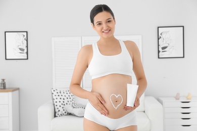 Photo of Pregnant woman with heart on her belly holding body cream at home