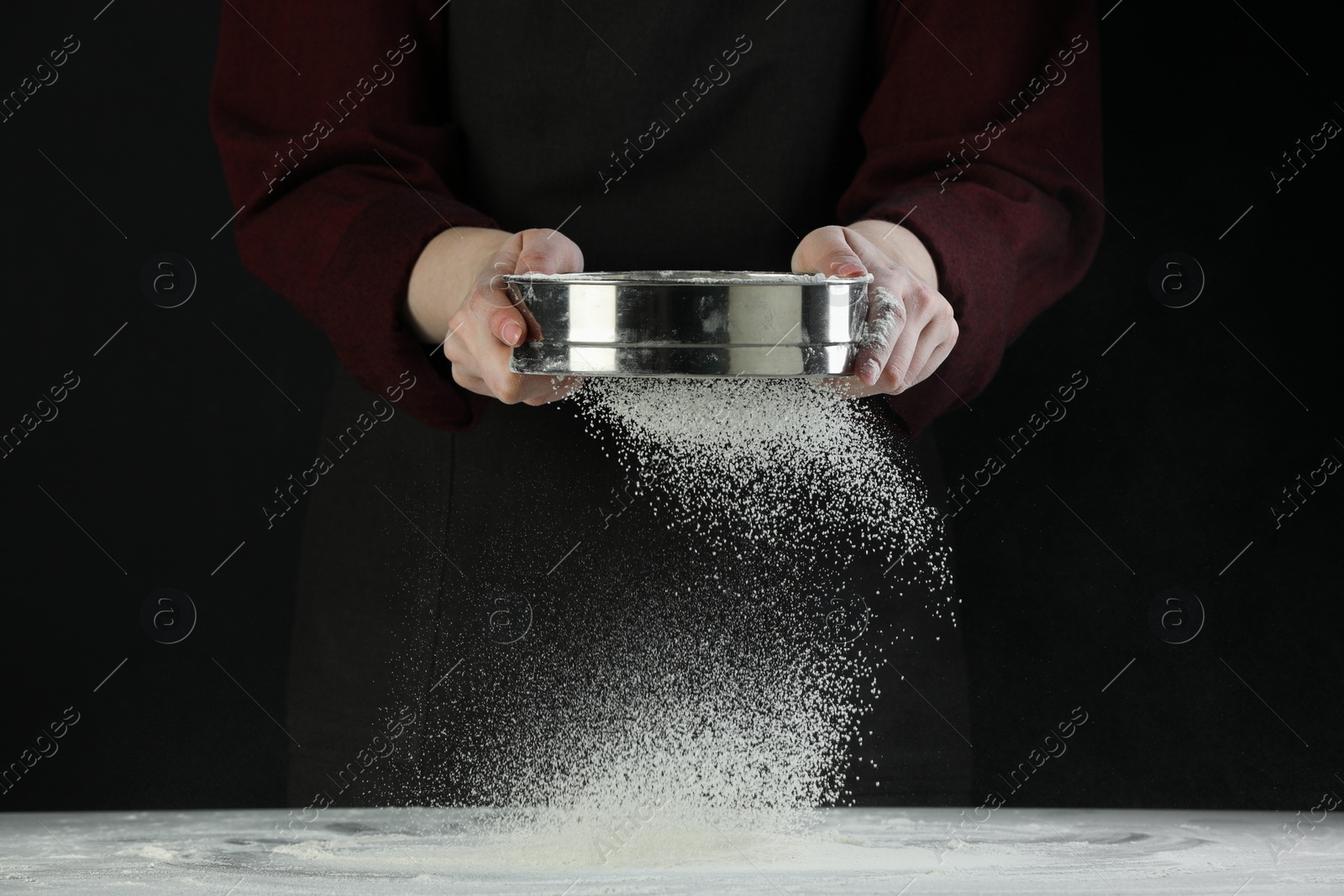 Photo of Woman sieving flour at table against black background, closeup