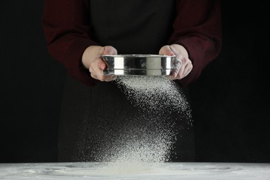 Woman sieving flour at table against black background, closeup