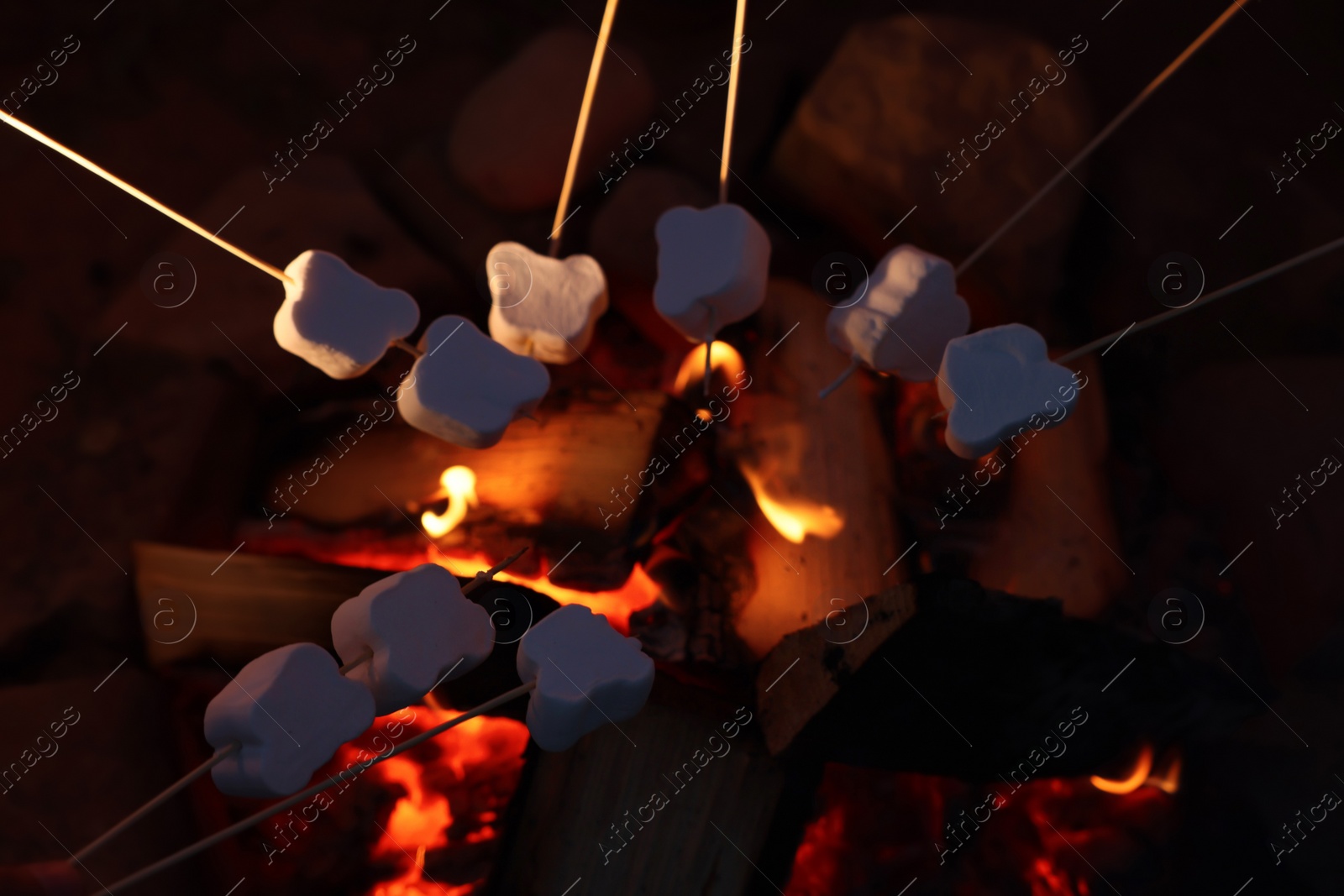 Photo of Delicious marshmallows roasting over bonfire outdoors at night, closeup. Camping season