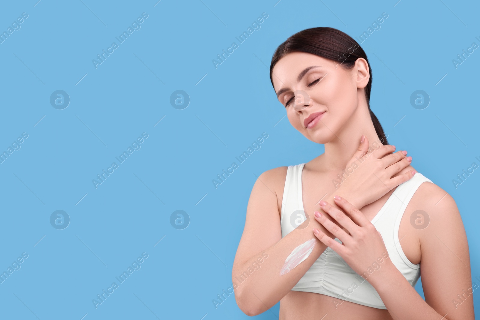 Photo of Beautiful woman with smear of body cream on her arm against light blue background, space for text