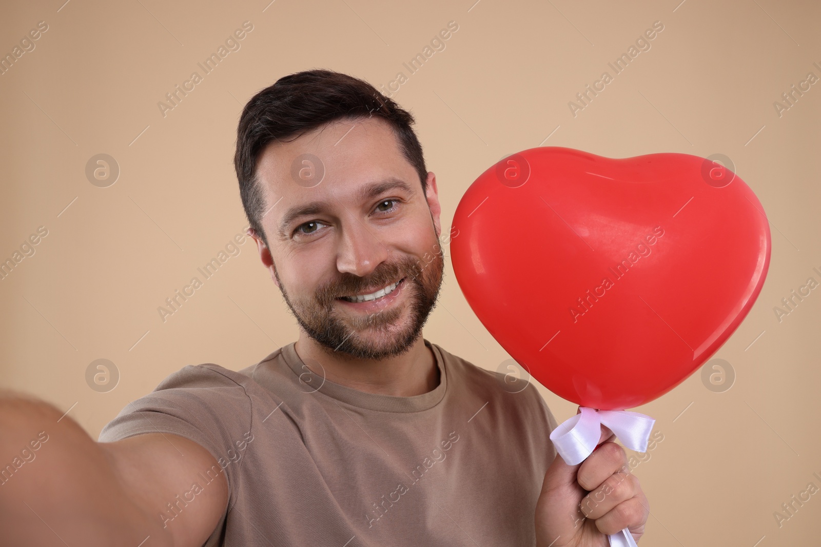 Photo of Man holding red heart shaped balloon and taking selfie on beige background