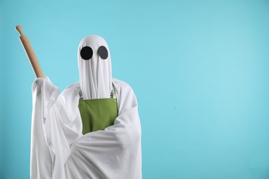 Woman in ghost costume and apron with rolling pin on light blue background, space for text