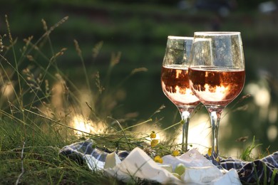 Glasses of delicious rose wine, cheese and grapes on picnic blanket near lake, space for text