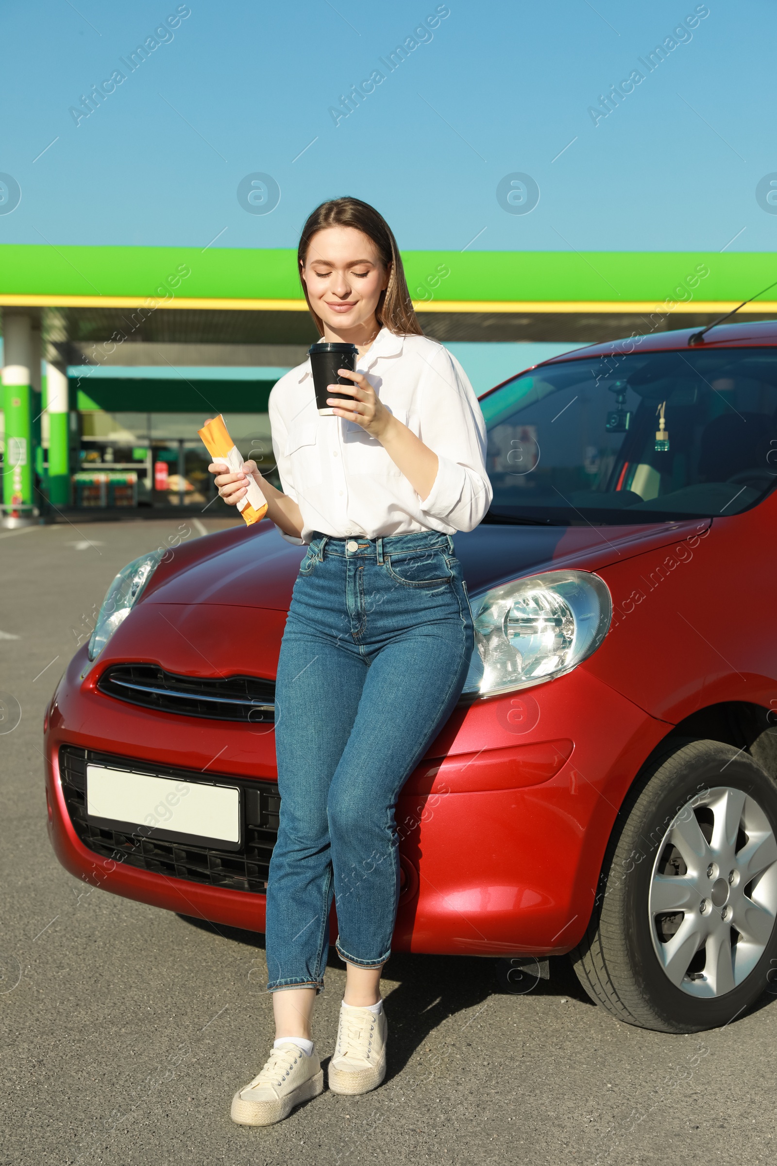 Photo of Beautiful young woman with hot dog drinking coffee near car at gas station