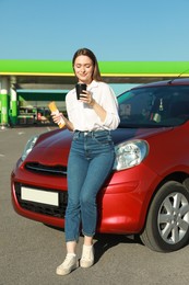 Photo of Beautiful young woman with hot dog drinking coffee near car at gas station