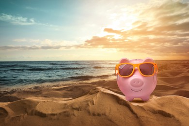Image of Saving money for summer vacation. Piggy bank with sunglasses on sandy beach near sea at sunset, space for text