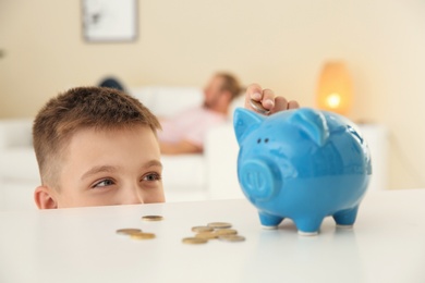 Photo of Little boy putting coin into piggy bank on table indoors