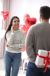 Photo of Young man presenting gift to his girlfriend in bedroom decorated with heart shaped balloons. Valentine's day celebration