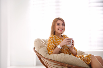 Photo of Young woman with cup of drink relaxing in papasan chair near window at home. Space for text