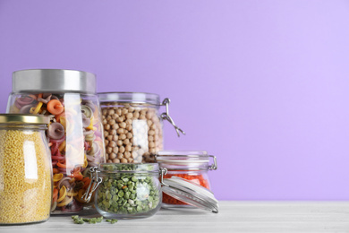 Photo of Jars with different cereals on white wooden table against violet background. Space for text