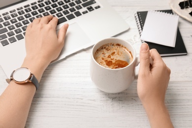 Photo of Young woman with cup of delicious coffee using laptop at table
