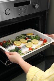 Photo of Woman putting baking dish with raw fish and vegetables into oven, closeup