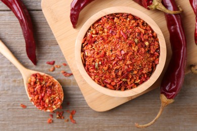 Photo of Aromatic spice. Red chili pepper flakes in bowl, spoon and pods on wooden table, flat lay