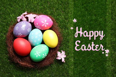 Image of Colorful eggs in decorative nest with flowers and text Happy Easter on green grass, top view